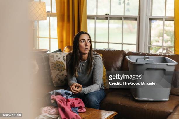 thoughtful woman looking away while sitting on sofa in living room - woman concerned stockfoto's en -beelden