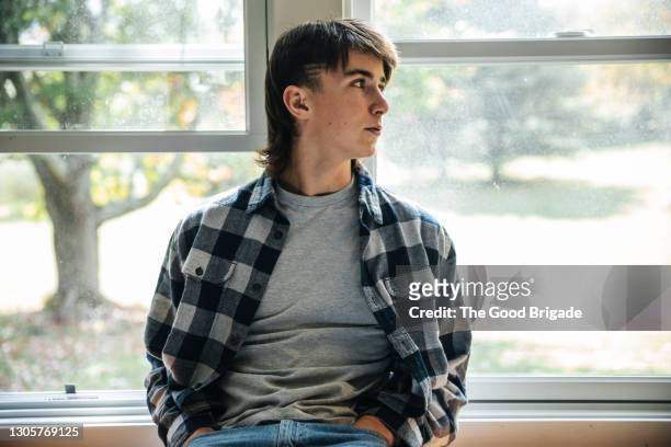 portrait of teenage boy looking away while sitting against window at home - mullet haircut stock pictures, royalty-free photos & images