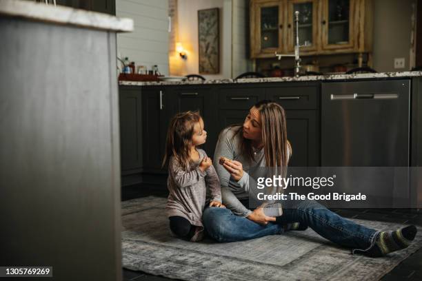 mother and young daughter sitting on kitchen floor at home - mother and child snacking stockfoto's en -beelden