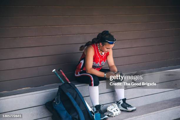 young female softball player looking at mobile phone in dugout - sports dugout fotografías e imágenes de stock
