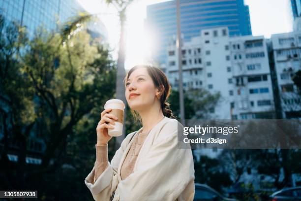 low angle portrait of confident and successful young asian businesswoman taking a coffee break in the middle of a work day, looking up to the sky and enjoying some fresh air, standing against corporate skyscrapers in the city - office wellbeing stock pictures, royalty-free photos & images