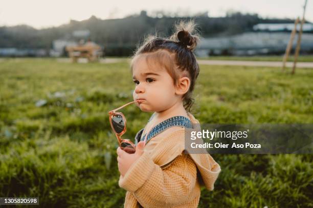 toddler looking left holding red little sun glasses and wearing denim pants and yellow jacket - baby sun hat stock-fotos und bilder