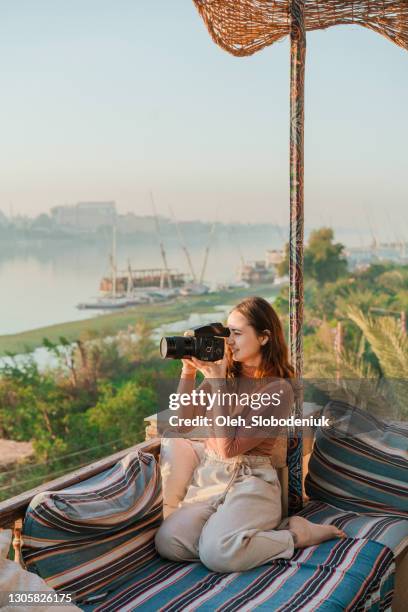 frau sitzt auf terrasse und fotograf nile bei sonnenuntergang in luxor - luxor stock-fotos und bilder