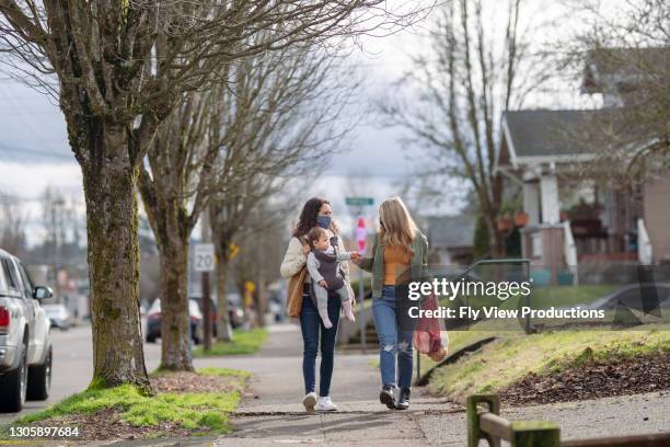 two women wearing protective face masks walking on residential sidewalk with baby girl - community safety stock pictures, royalty-free photos & images