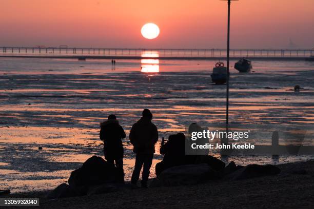 People sit on the beach and watch the sunset over the pier on February 27, 2021 in Southend-on-Sea, England.