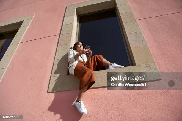 young woman with smart phone sitting on window sill of building - window sill stock pictures, royalty-free photos & images