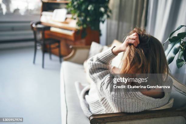 a woman with her back to the camera is lying on a sofa in a beautiful room with a piano, her hand holding her head. depression, illness, imprisonment, boredom, loneliness. - senza speranza foto e immagini stock