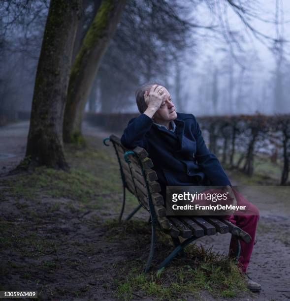 holding hand on his head, a senior man sits on a bench alone in misty forest, lonely and abandoned in gloomy atmospheric mood - bench stock pictures, royalty-free photos & images