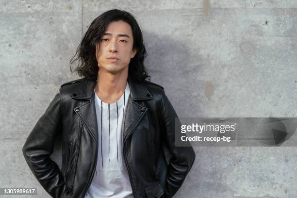 portrait of cool young handsome man wearing leather jacket and leaning against concrete wall - long hair stock pictures, royalty-free photos & images