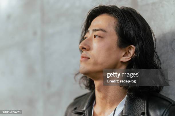 portrait of cool young handsome man wearing leather jacket and leaning against concrete wall - asain model men stock pictures, royalty-free photos & images