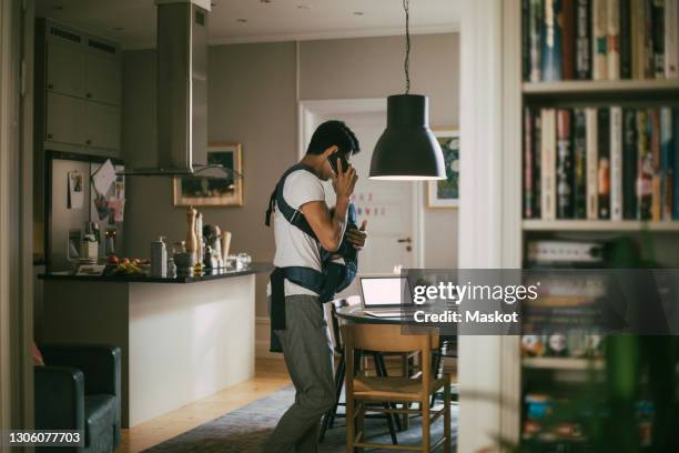 father talking on smart phone while carrying male toddler in kitchen - stay at home saying stock pictures, royalty-free photos & images