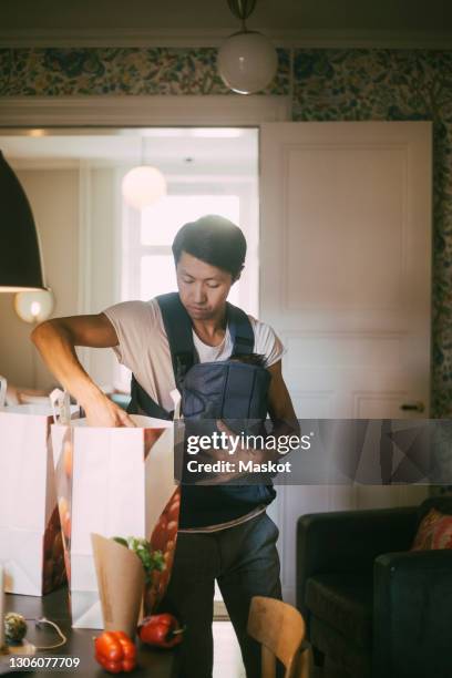 father carrying baby boy while looking in grocery bag at home - baby care stock pictures, royalty-free photos & images