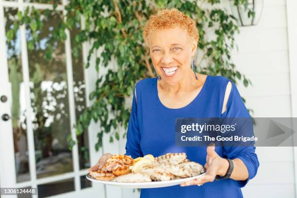 portrait of smiling senior woman holding platter of food on patio - meat platter stock-fotos und bilder