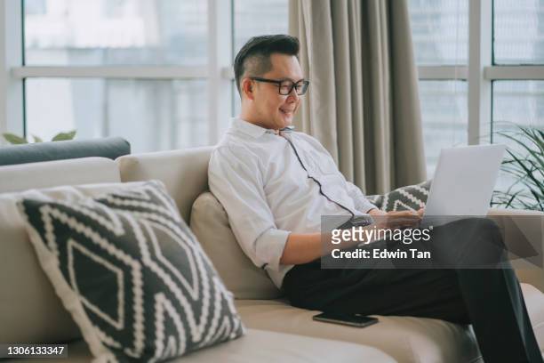 asiatische chinesische mann sitzt auf seinem sofa mit kissen entspannen donerisch mit seinem laptop surfen im netz und online-shopping - airport asian worker stock-fotos und bilder