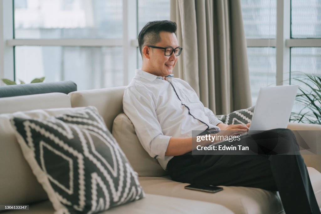 Asian chinese man sitting on his sofa with cushion relaxing with his laptop surfing the net and shopping online