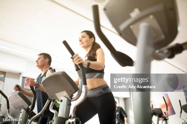 attraktive 40-jährige frau führt elliptisches fahrradtraining in einem fitnessstudio mit einem reifen partner im hintergrund durch - cross trainer stock-fotos und bilder