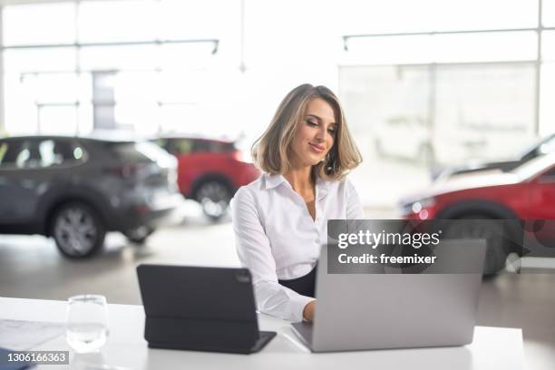 jonge elegante verkoopvrouwzitting in de saloon van de autodealer en het gebruiken van laptop - car salesman stockfoto's en -beelden