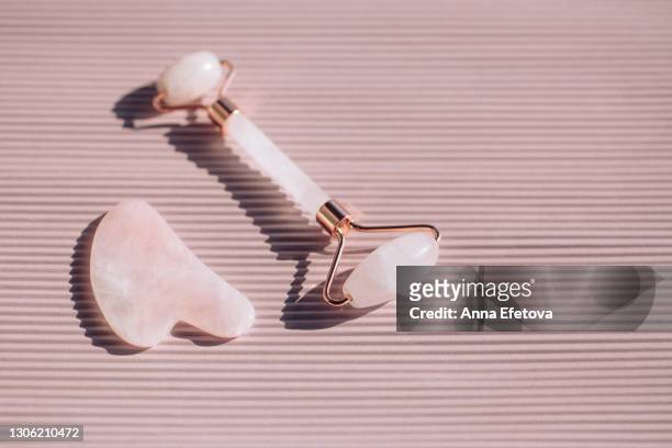 rose quartz roller and guasha on striped pastel light pink background. massagers are used for anti aging procedures with an oil or moisturizing lotion. sunlight makes shadows and illuminating reflections from tools. flat lay style with copy space - jade gemstone stock pictures, royalty-free photos & images
