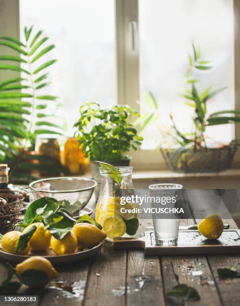 glass jug with fresh lemon and glass of lemonade on wooden kitchen table with ingredients at window. - cocktails water stock pictures, royalty-free photos & images