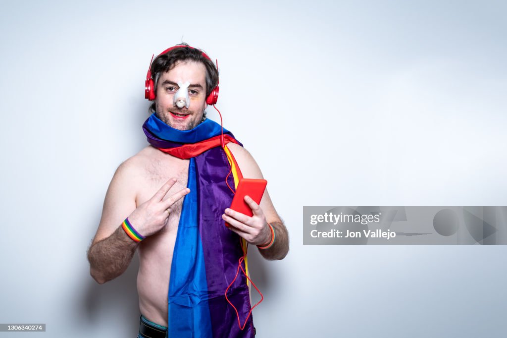 A gay man with red lipstick and a broken nose and stitches is with the LGTBI flag wrapped around his neck. He is looking straight ahead smiling and is listening to music with red headphones that match the cell phone case he is holding in his hand.