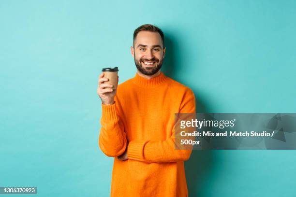 portrait of smiling young man holding coffee cup while standing against blue background - hipster beard plain background stock-fotos und bilder