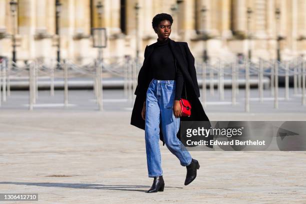 Model wears a black turtleneck wool pullover, a wool black long coat, blue denim jeans, black leather pointy boots, a red bag, outside Vuitton, on...