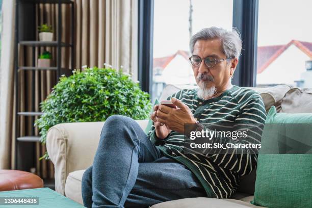portrait of asian senior man using smart phone while sitting on sofa in living room at home, senior man typing an sms message and working mobile devices, people lifestyle technology concept - older indian man stock pictures, royalty-free photos & images