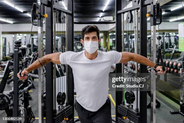 young man using apparatus during strength training at the gym with face mask - gym reopening stock pictures, royalty-free photos & images