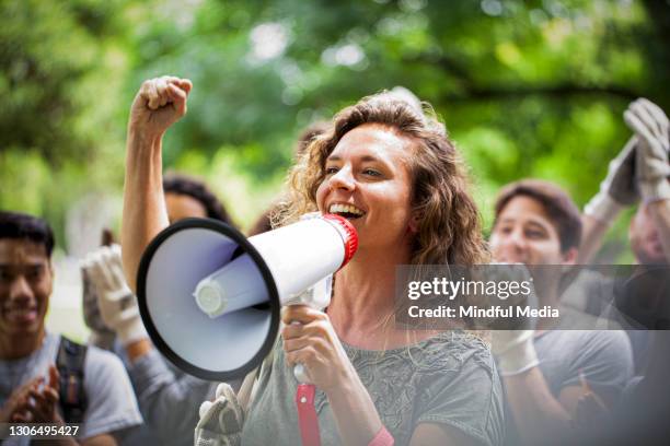 young woman activist holding megaphone and giving a speech with her fist up during garbage cleanup event - eco activist stock pictures, royalty-free photos & images