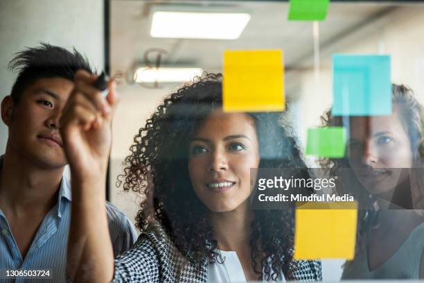 smiling businesswomen and businessman looking at adhesive notes while writing on glass wall - three people stock pictures, royalty-free photos & images
