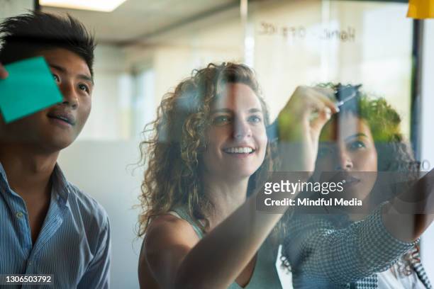 smiling businesswomen and businessman looking at adhesive notes while writing on glass wall - creative director stock pictures, royalty-free photos & images