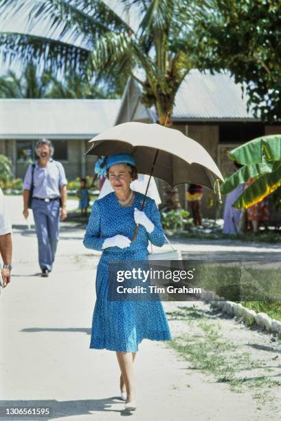 British Royal Queen Elizabeth II, wearing a blue two-piece outfit with white detail and a matching hat, shades herself beneath a parasol during a...