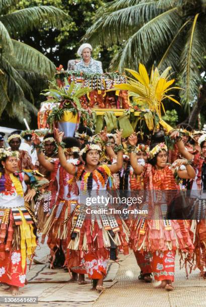 British Royal Queen Elizabeth ll, wearing a floral print outfit with a hat designed by milliner Frederick Fox, carried on a local canoe by Tuvaluans...