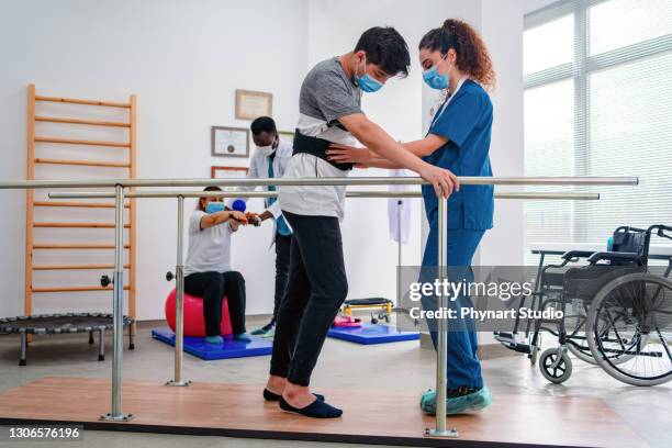 jonge mens die parallelle staven gebruikt om te lopen en fysiotherapeut die hem helpt terwijl een andere patiënt en professional op de achtergrond lopen - afkickcentrum stockfoto's en -beelden