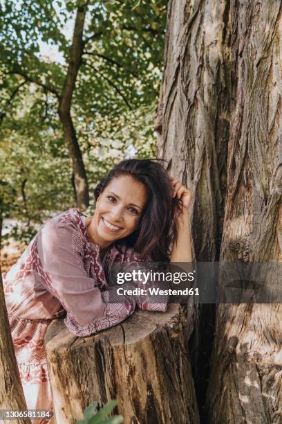 smiling woman with hand in hair resting on tree trunk at park - hand in hair stock pictures, royalty-free photos & images