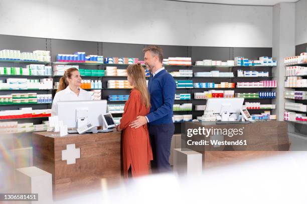 Couple Talking To Female Pharmacist At Pharmacy Checkout, Stockfoto