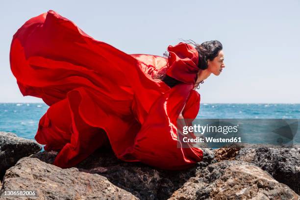 confident female dancer in flowy red dress on rocks by seashore during sunny day - woman long dress beach stock pictures, royalty-free photos & images