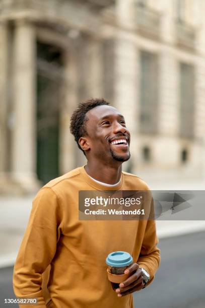 smiling young man with disposable cup looking away while standing in city - coffe to go stock pictures, royalty-free photos & images