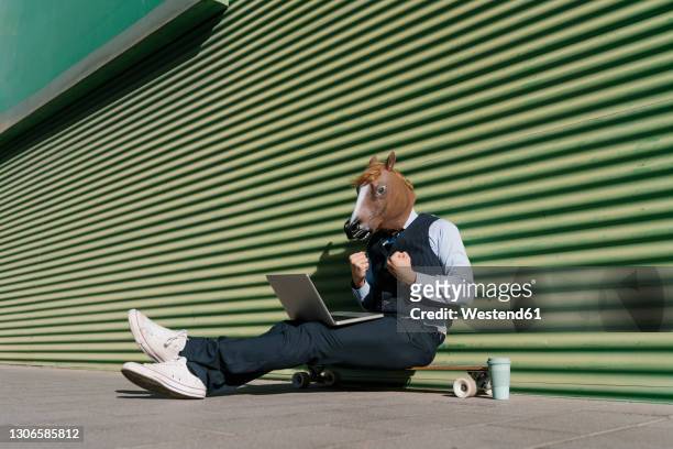 businessman in horse mask with laptop gesturing while sitting against green wall - crazy man computer stock-fotos und bilder