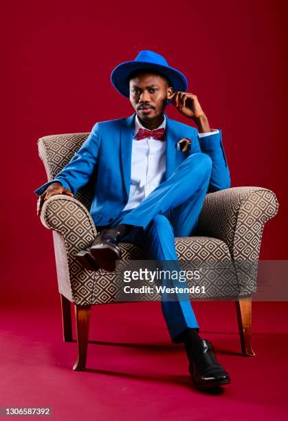 confident man wearing blue suit sitting on chair against red background - red suit stockfoto's en -beelden