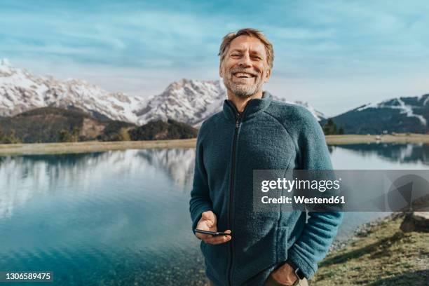 happy mature man holding mobile phone while standing against prinzensee, salzburger land, austria - tourist guy stock pictures, royalty-free photos & images