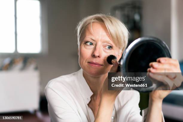 mature woman doing make-up while looking at mirror - blusher stock-fotos und bilder