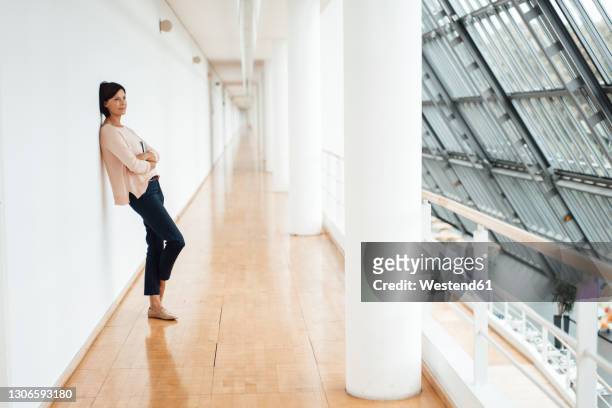 businesswoman with arms crossed leaning against wall on corridor at office - leaning stock pictures, royalty-free photos & images
