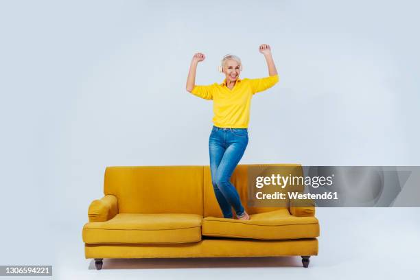 cheerful senior woman dancing on sofa while listening music through wireless headphones against white background - senior woman studio ストックフォトと画像