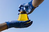 Hand with gloves holding beaker with ethanol biofuel against blue sky