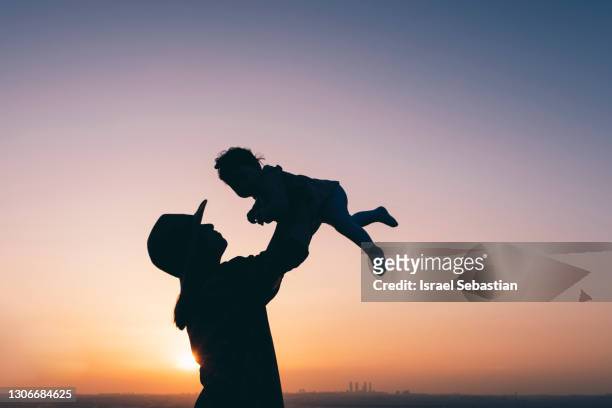 silhouette of a mother with a hat holding her baby up in the air in a beautiful sunset. - short phrase stock pictures, royalty-free photos & images
