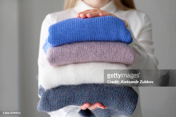 a woman or a girl holds an armful of knitted things of different colors, stacked in a pile, in the room. winter and autumn warm cozy sweaters for charity. the concept of storage, care and washing of handmade products. a copy of the text space. - winter fashion collection stock-fotos und bilder
