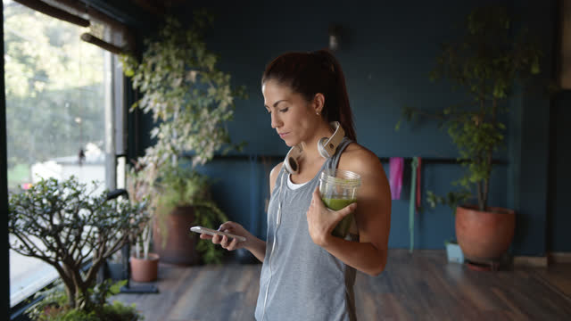 Young Latin American woman drinking a green juice at the gym while checking messages on smartphone with headphones around her neck