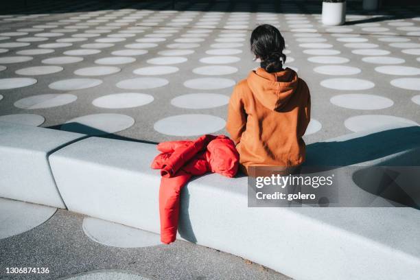 girl in orange hoody sitting on stone bench on white spotted town square - modern town square stock pictures, royalty-free photos & images