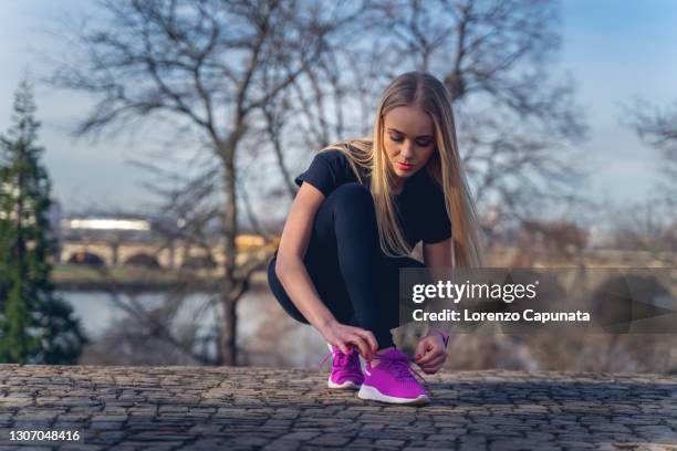 young woman, in a park, illuminated by sunlight, lowered down, fastening her running shoes, ready to train. - lace fastener stock-fotos und bilder